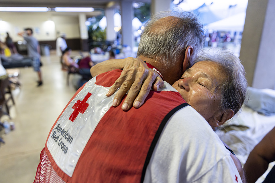 August 14, 2023. Wailuku, Hawaii.
American Red Cross volunteer Ned Worman comforts Elaine Goode at the Red Cross shelter at the War Memorial Gymnasium in Wailuku on Maui. Goode lost everything when a wildfire destroyed her home in Lahaina. Among the irreplaceable items she lost in the fire were the ashes of her recently deceased husband. 

“There are things you can't even replace,” Goode said as tears filled her eyes. “I don't even have a picture of him. I don't have his picture. I had a picture of us when we first got together 32 years ago. And I don't have anything. I don't have his urn.”

The Lahaina wildfire was the deadliest fire in the United States in more than 100 years. It and destroyed as many as 2,700 structures, most of which were homes, leaving thousands of residents without somewhere to live.
Photo by Scott Dalton/American Red Cross