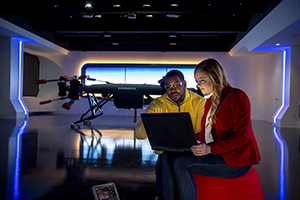 A picture of a Man and Woman with safety glasses on looking at laptop screen with large UAV quad-rotor in background.