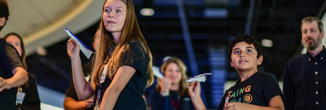 Multiple school children holding paper airplanes, facing the camera, reading their planes to fly