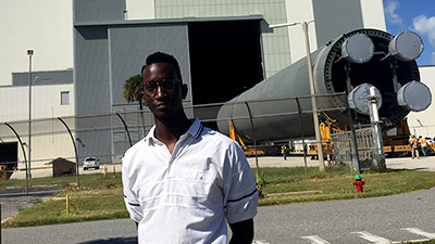 Brandon Burroughs standing in front of Kennedy Space Center Vehicle Assembly building with Space Launch System pathfinder.