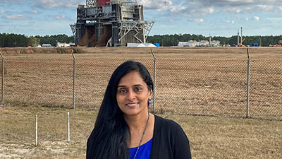 Boeing employee Suba Iyer stands outdoors with a rocket test stand in the background. 